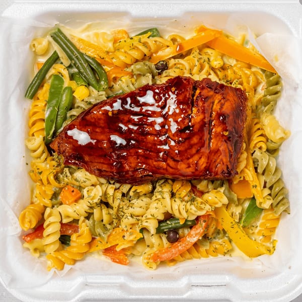 A takeout container of Jamaican jerk salmon over pasta with colorful vegetables and creamy sauce.